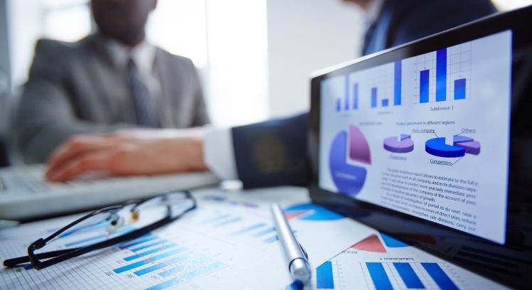 Image of eyeglasses, pen, touchpad and financial documents at workplace with businessmen working on background